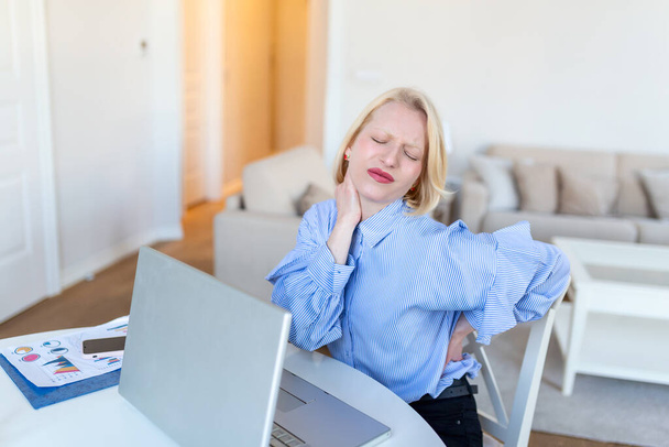 Portrait of young stressed woman sitting at home office desk in front of laptop, touching aching back with pained expression, suffering from backache after working on laptop - Foto, Imagem