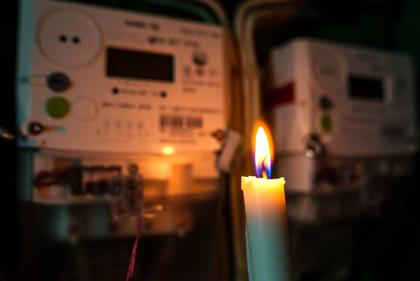 火のろうそくの光に照らされた電気メーター。停電、エネルギー危機または停電の概念イメージ. - 写真・画像