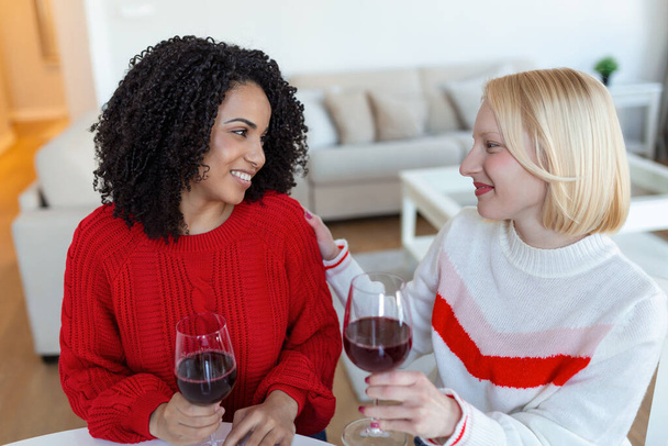 Naiset istuvat sohvalla nauraen viihtyisässä asunnossa viinin kanssa. Kaksi naispuolista ystävää rentoutumassa sohvalla kotona lasin kanssa viiniä puhumassa yhdessä. - Valokuva, kuva