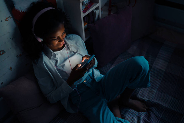 African smiling teen girl sitting on a bed with phone texting or using mobile apps in her room at home - Fotoğraf, Görsel