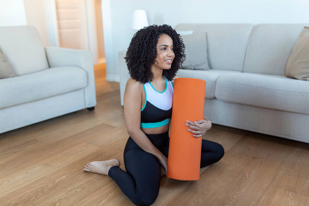 Beautiful young African woman in sports wear is holding a yoga mat and a bottle of water, looking away and smiling, standing at home - Photo, Image