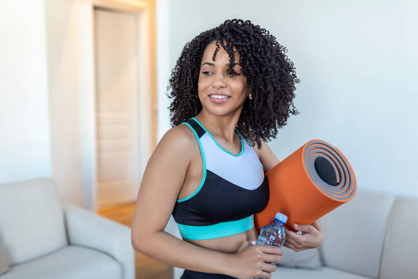 Beautiful young African woman in sports wear is holding a yoga mat and a bottle of water, looking away and smiling, standing at home - Foto, Imagen