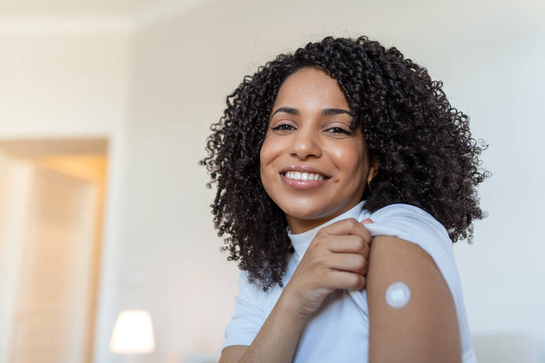 Portrait of a female smiling after getting a vaccine. Woman holding down her shirt sleeve and showing her arm with bandage after receiving vaccination. - Foto, afbeelding