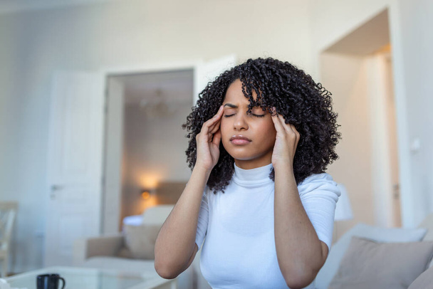 Portrait of an attractive woman sitting on a sofa at home with a headache, feeling pain and with an expression of being unwell. Upset depressed woman lying on couch feeling strong headache migraine. - Photo, Image