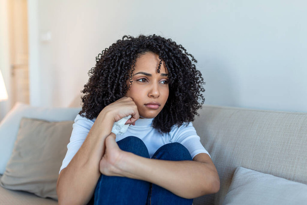 Νεαρή Αφρο-Αμερικανίδα που νιώθει αναστατωμένη, λυπημένη, δυστυχισμένη ή απογοητευμένη κλαίγοντας μόνη στο δωμάτιό της. Γυναίκα που υποφέρει από κατάθλιψη κάθεται στο κρεβάτι και κλαίει - Φωτογραφία, εικόνα