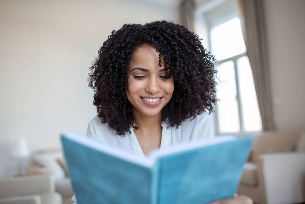 Young beautiful African American woman holding book, University student studying, learning language. leisure, literature and people concept - smiling african american woman reading book at home - Fotó, kép