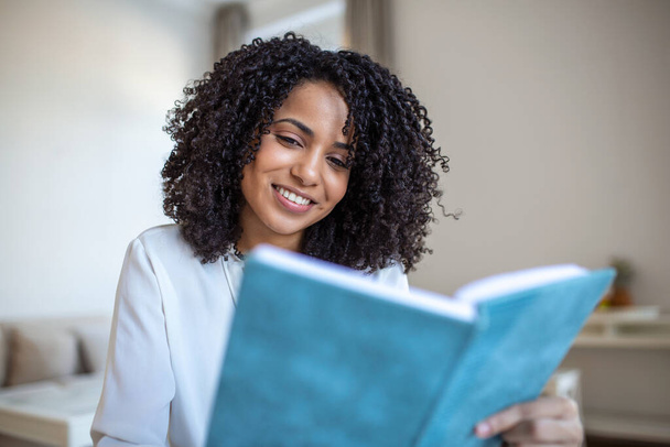Young beautiful African American woman holding book, University student studying, learning language. leisure, literature and people concept - smiling african american woman reading book at home - Fotó, kép