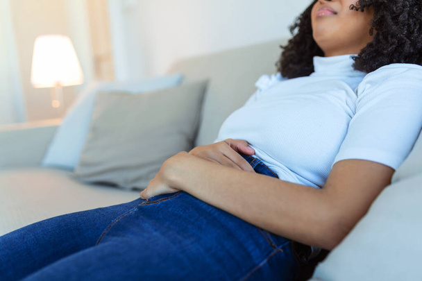 African American Woman in painful expression holding hands against belly suffering menstrual period pain, lying sad on home bed, having tummy cramp in female health concept - Photo, Image