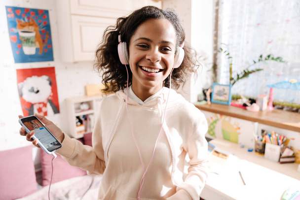 African happy teen in headphones listening to music and dancing in her cozy room at home - Photo, Image