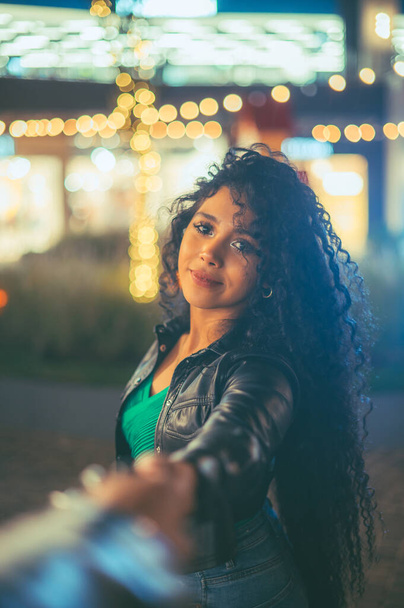 Teenage girl with curly hair, of Latin origin, walking in a shopping center full of lights, wearing a leather jacket and a green blouse - Φωτογραφία, εικόνα