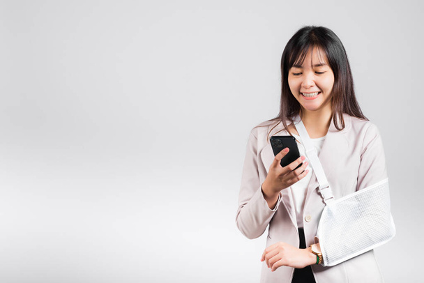 Mujer confiada sonriendo brazo roto después del accidente y usar férula del brazo para el tratamiento y mantener el teléfono inteligente, la honda asiática feliz mano apoyada aislada sobre fondo blanco, redes sociales, espacio de copia - Foto, imagen
