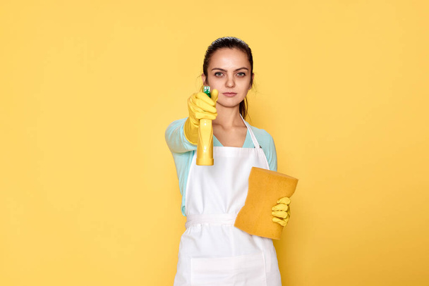 caucasian woman in yellow rubber gloves and cleaner apron with sponge and detergent sprayer on yellow background. - Foto, immagini
