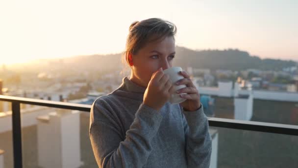 Frau beginnt ihren Tag mit einer Tasse Tee oder Kaffee auf dem Balkon im Morgengrauen, Zeitlupe. Stadtlandschaft im Hintergrund. Moderner urbaner Lebensstil - Filmmaterial, Video