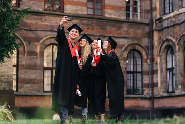 grupo de estudiantes felices en batas de soltero con diplomas tomando una selfie en el teléfono al aire libre. - Foto, imagen