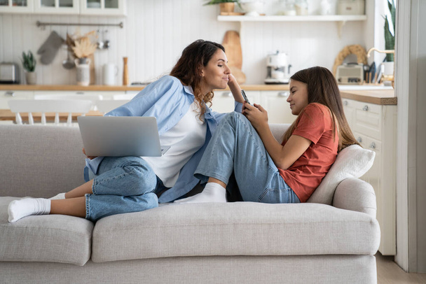  Young smiling woman sitting on sofa with laptop spying on child teen girl using cellphone, mom and kid spending leisure time together on devices. Parental control and smartphone generation - Фото, изображение