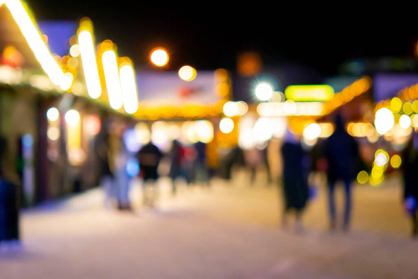 Fond flou. Les gens marchent sur la place de la ville la nuit d'hiver. Silhouettes noires de personnes marchant près des maisons décorées illumination lumineuse. Lumière blanche taches de flou bokeh de lumières de maison éclatantes - Photo, image