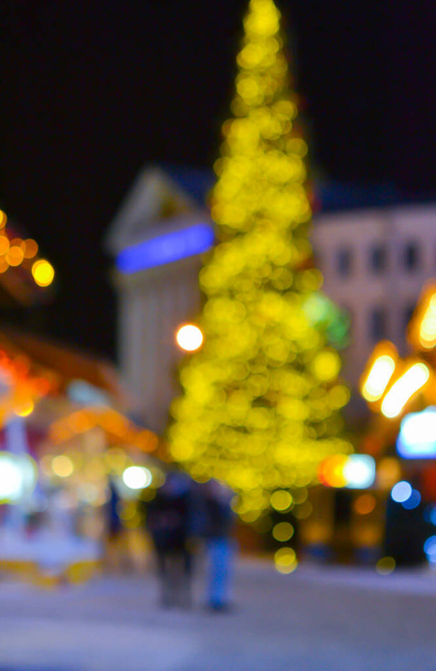 Arka plan bulanık. Noel ağacı, dekore edilmiş parlak ışıklar, bina kış gecesi şehir meydanında yürüyen insanların bulanık siluetleri. Güzel yeni yıl ve Noel tatili geçmişi bulanıklaştırıyor - Fotoğraf, Görsel