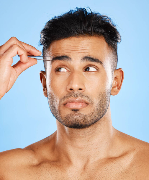 Face, hair removal and man in studio with tweezer for skincare, beauty and eyebrow on blue background. Tweezing, wellness and indian model shape eyebrows for grooming, hygiene and cleaning on mockup. - Photo, Image