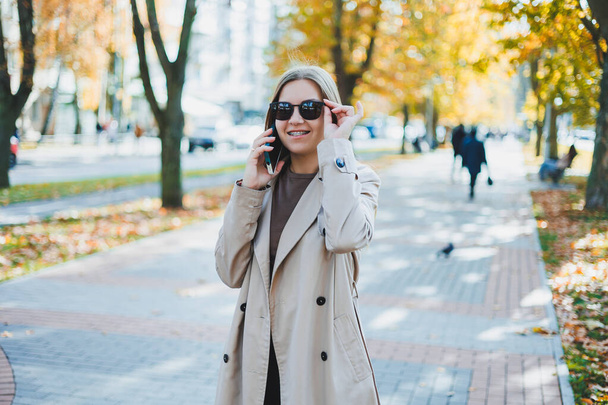 A beautiful woman in sunglasses is walking in the autumn park and talking on the phone. Joyful girl on the autumn alley in the city. - Fotografie, Obrázek