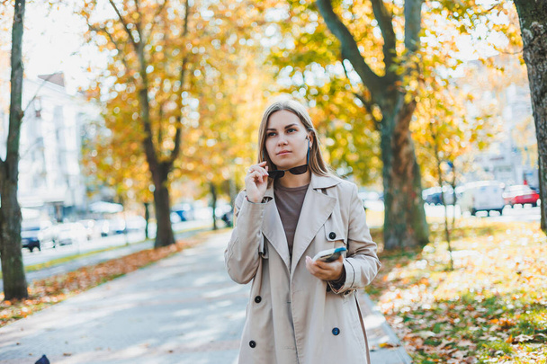 Güneş gözlüklü güzel bir kadın sonbahar parkında yürüyor ve telefonda konuşuyor. Şehrin sonbahar sokağında neşeli bir kız. - Fotoğraf, Görsel