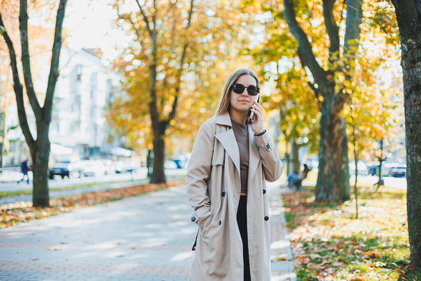 Young smiling woman walking in autumn park, talking on smartphone, wearing trench coat, sunglasses, happy mood, fashion style trend - Photo, Image