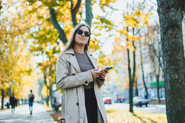 Стильная счастливая женщина в осеннем пальто и солнцезащитных очках гуляет на улице и разговаривает по мобильному телефону, веселая женщина разговаривает и болтает по мобильному телефону на открытом воздухе во время прогулки в парке - Фото, изображение