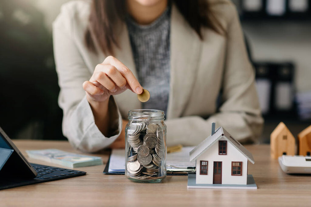 woman putting money coins into a glass bottle jar. wooden house small model on table - Photo, Image