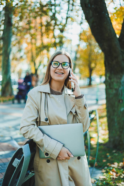 Красива молода жінка в осінньому пальто, використовуючи ноутбук і телефон, працює віддалено, жінка розмовляє на мобільному телефоні в осінньому парку
. - Фото, зображення