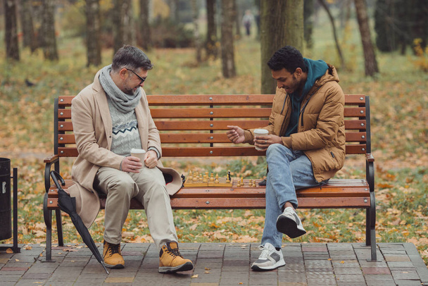 Uomini multietnici, insegnante e studente che giocano a scacchi nel parco autunnale su una panchina. - Foto, immagini