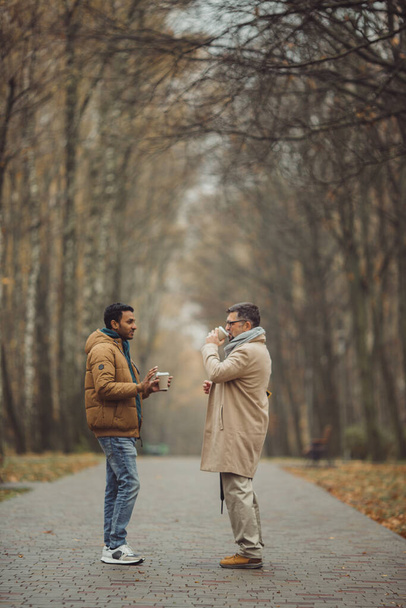 Friends, a senior and a young man walking and talking and drinking coffee together in the autumn park - Фото, изображение