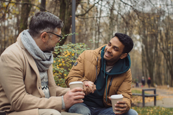 Χαμογελώντας πολυεθνικοί άνδρες που μιλούν στον πάγκο στο πάρκο το φθινόπωρο - Φωτογραφία, εικόνα