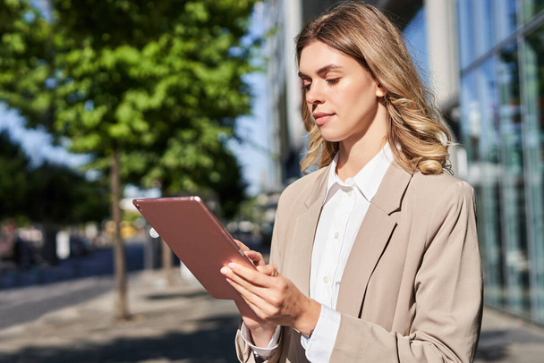 Portrait of corporate woman reads news, works on her digital tablet while on her way to office, stansd on street of city center in suit. - Foto, Imagem