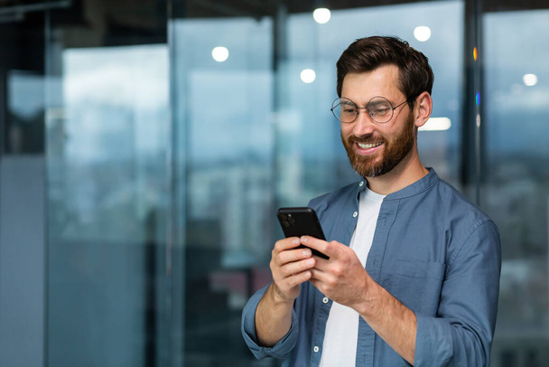 Successful businessman in glasses and beard standing near window in the evening, man using phone, happy boss typing message and browsing online pages on smartphone app inside office. - Foto, Bild