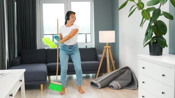 Mulher branca em fones de ouvido limpando a casa e se divertindo dançando com uma vassoura e pano. Movimento lento - Filmagem, Vídeo