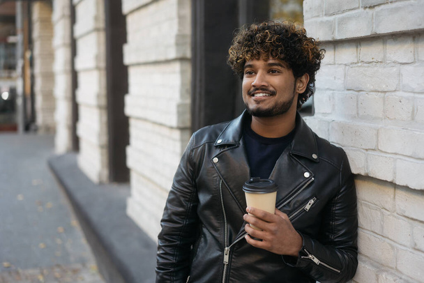 Jóképű, mosolygós indiai férfi fekete bőrdzsekiben, kezében egy csésze kávéval, miközben az utcára néz. Portré elegáns ázsiai hipster áll fal mellett forró ital, másolás tér  - Fotó, kép
