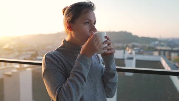 A nő egy csésze teával vagy kávéval kezdi a napját az erkélyen hajnalban, lassítva. Városi táj a háttérben. Modern városi életmód - Felvétel, videó
