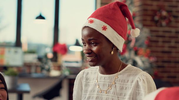 アフリカ系アメリカ人女性は、冬のイベント中にオフィスパーティーの同僚とクリスマス前夜を祝う。休日のお祝いで楽しいサンタの帽子を持つお祭りの人。手持ち撮影. - 写真・画像