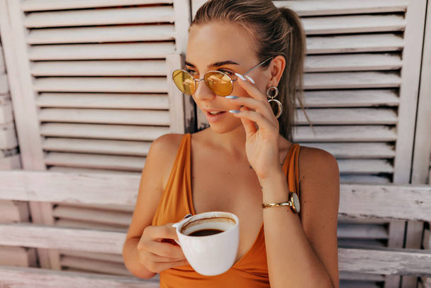 Close up outdoor portrait of romantic pretty woman with collected hair wearing round orange glasses and orange t-shirt sitting outdoor with coffee and looking aside. - Foto, afbeelding