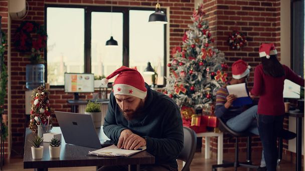 Kierownik projektu pracujący na laptopie w uroczystym biurze z ozdobami świątecznymi, wykonujący pracę raportów startupowych w okresie ferii zimowych. Człowiek wysyłając e-mail w przestrzeni z lampkami świątecznymi. - Zdjęcie, obraz