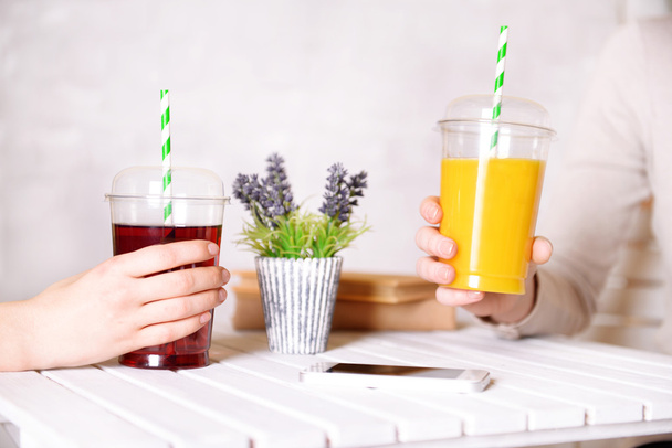 Two female hands with fast food cups of juice at wooden table with plant on light wall background - Fotografie, Obrázek