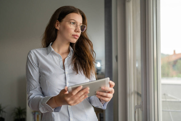 One woman young adult caucasian female stand at her the window at work or home front view waist up copy space wear shirt looking confident with eyeglasses hold digital tablet - Foto, afbeelding