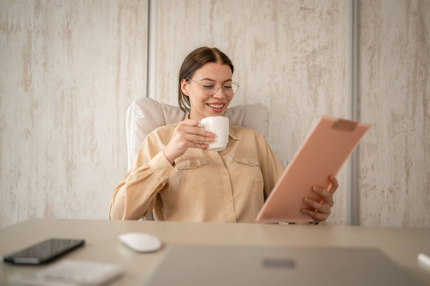 one woman young adult caucasian female secretary or manager or worker sit at the table at her office desk taking a brake from work real people copy space happy smile hold cup of coffee - Photo, Image