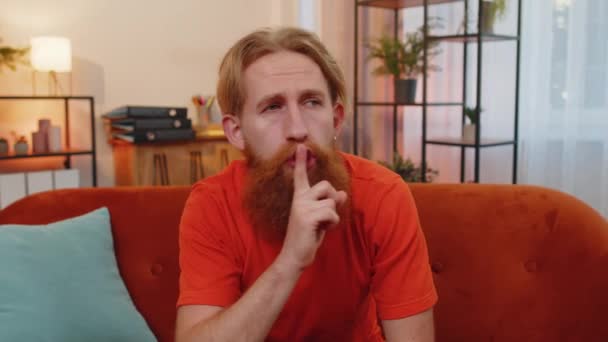 Shh be quiet, hush please. Bearded redhead man presses index finger to lips makes silence hush gesture sign do not tells gossip secret. Young guy at home apartment living room sitting on orange couch - Footage, Video