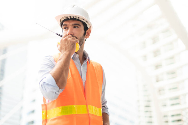 Beyaz adam mühendisleri konuşmak için telsiz kullanırlar. Şehir merkezindeki inşaat alanında turuncu yelek ve büyük başlıklar giyerler.. - Fotoğraf, Görsel
