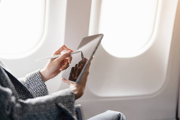 Een succesvolle Aziatische zakenvrouw of vrouwelijke ondernemer in formele pak in een vliegtuig zit in een business classs stoel en maakt gebruik van een tablet computer tijdens de vlucht. - Foto, afbeelding