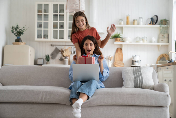 Cheerful overjoyed woman sits on sofa holding gift box from teenage daughter. Satisfied European girl smiles and makes present surprise standing behind mother for concept happy family and motherhood - Photo, Image