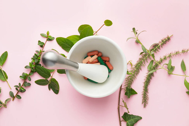 Mortar with pills and different herbs on pink background - Photo, image