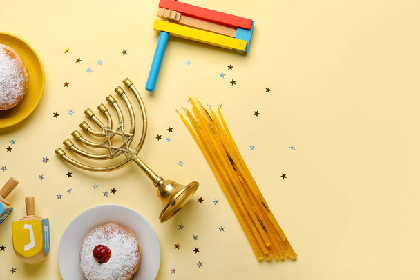 Plates with donuts, menorah, dreidels, candles and rattle for Hanukkah celebration on yellow background - Fotó, kép