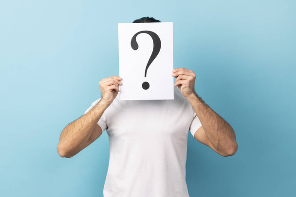 Portrait of man wearing white T-shirt hiding his face behind white paper with question mark, finding smart solution, asking for advice. Indoor studio shot isolated on blue background. - Foto, Bild