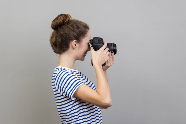 Side view of woman photographer or traveler wearing striped T-shirt holding professional digital dslr camera and focusing, taking photo, shooting video. Indoor studio shot isolated on gray background. - Photo, Image